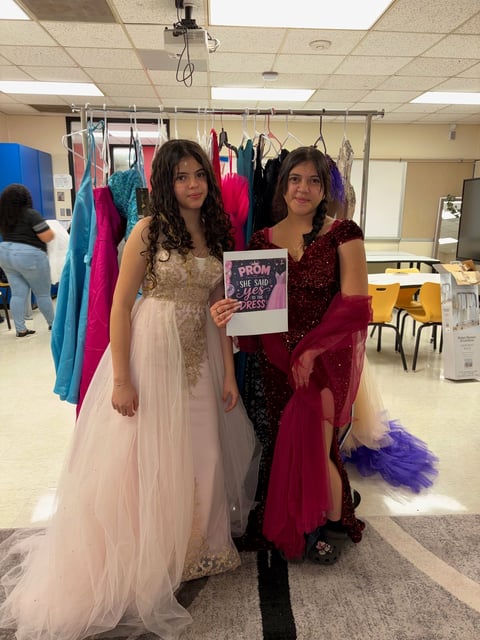 Two teenage girls in formal prom dresses holding a Prom sign in a school gymnasium with clothes racks behind them