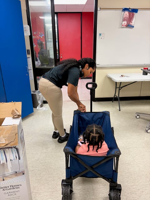 Woman bending over a blue stroller with a child inside in an indoor classroom or office setting