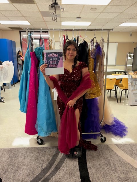 Woman in burgundy sequin dress holds sign promoting prom dress in clothing boutique with colorful gowns on rack