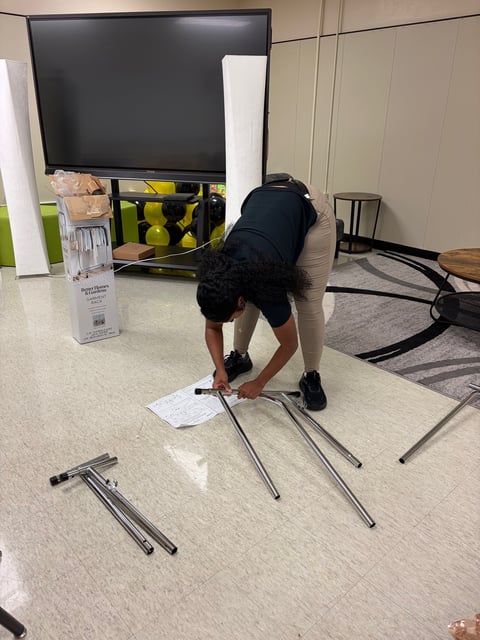 Person bending over metal leg frames on floor with TV and gym equipment in background