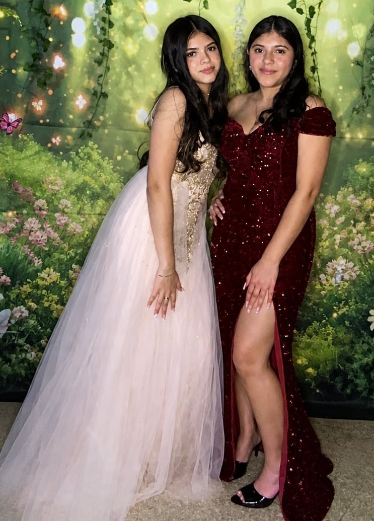 Two young women posing together at a formal event with an illuminated green floral backdrop with hanging vines and warm lights