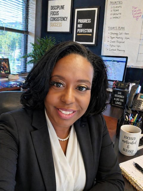Professional woman in black blazer and white blouse smiling at desk in office with motivational signs and computer visible in background
