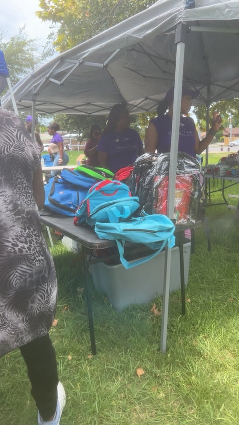 Outdoor event tent with vendor booth displaying colorful backpacks and merchandise in a grassy park setting