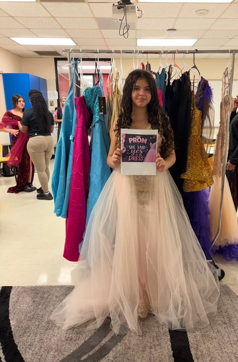 Young woman modeling a white tulle prom dress in a dressing room holding a sign that says Prom Dress, with colorful gowns hanging in background