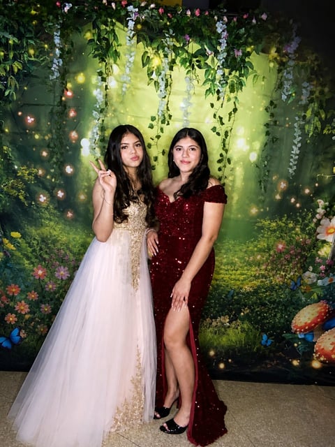 Two young women in formal dresses posing together in front of a lush green floral backdrop with glowing lights